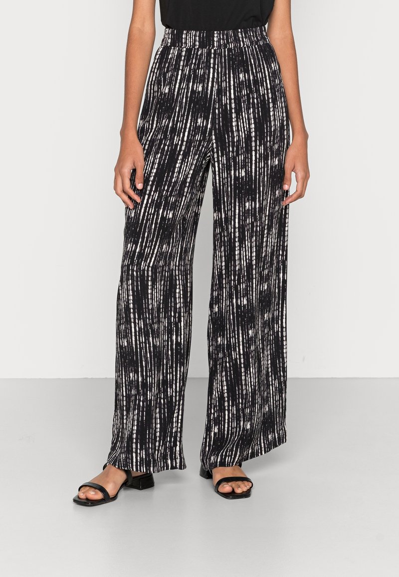 Femme portant un pantalon large à rayures verticales noires et blanches avec une taille élastique et des sandales noires à bout ouvert.