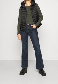 Svart pufferväst med huva, ihop med en olivgrön långärmad topp och mörkblå utsvängda jeans. Kraftiga svarta stövlar fulländar looken.