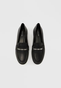 Μαύρα δερμάτινα μοκασίνια με λείο φινίρισμα, διακοσμημένα με ασημένια αλυσίδα στο μπροστινό μέρος και σχεδιασμό με στρογγυλεμένη μύτη.