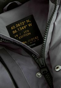Intérieur de la veste grise présentant une étiquette en tissu avec des coordonnées, un numéro d'édition et des détails de fabrication, ainsi qu'une fermeture éclair noire.