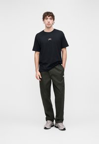 T-shirt preta de algodão com um pequeno logótipo impresso, combinada com calças cargo verdes com bolsos laterais, e sapatilhas cinza claro.