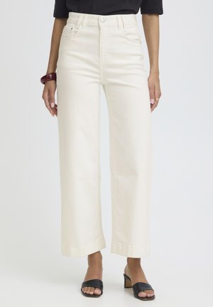 Frau trägt hoch taillierte, weit geschnittene, off-white Hose, schwarze Sandaletten mit Absatz, schwarzes Oberteil und ein dunkelrotes Armband am linken Handgelenk.