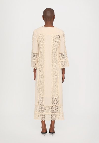 Beige dress with intricate lace details along the sleeves and hem. Flared sleeves, knee-length, simple round neckline, lightweight fabric.