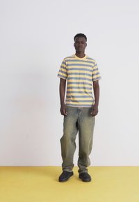 Camiseta a rayas en amarillo y gris, combinada con jeans holgados de lavado claro y calzado negro, sobre un fondo blanco con un suelo amarillo.