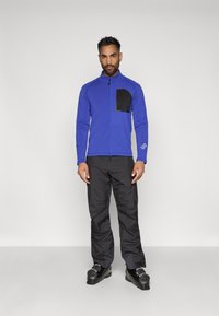 Plavi flis jakna s crnom prsnom džepom, u kombinaciji s crnim hlačama i crnim cipelama. Model stoji protiv prazne bijele pozadine.