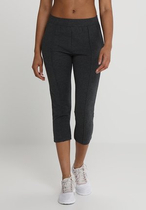 Femme portant des leggings courts gris foncé et des baskets blanches, debout devant un fond clair uni.