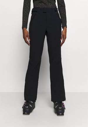 Pantalon de ski noir porté avec des chaussures de ski noires et un haut à manches longues à motifs, vu du milieu du torse jusqu’aux pieds sur un fond uni.