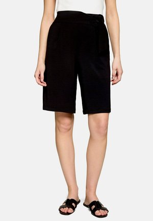 Femme portant un short noir au genou avec une ceinture à nouer, un haut blanc sans manches et des sandales plates noires à bout ouvert, debout devant un fond uni.