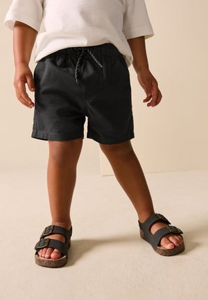 Kind dat zwarte shorts met trekkoord, een wit shirt met korte mouwen en zwarte sandalen met gesp draagt, staand op een lichte vloer.