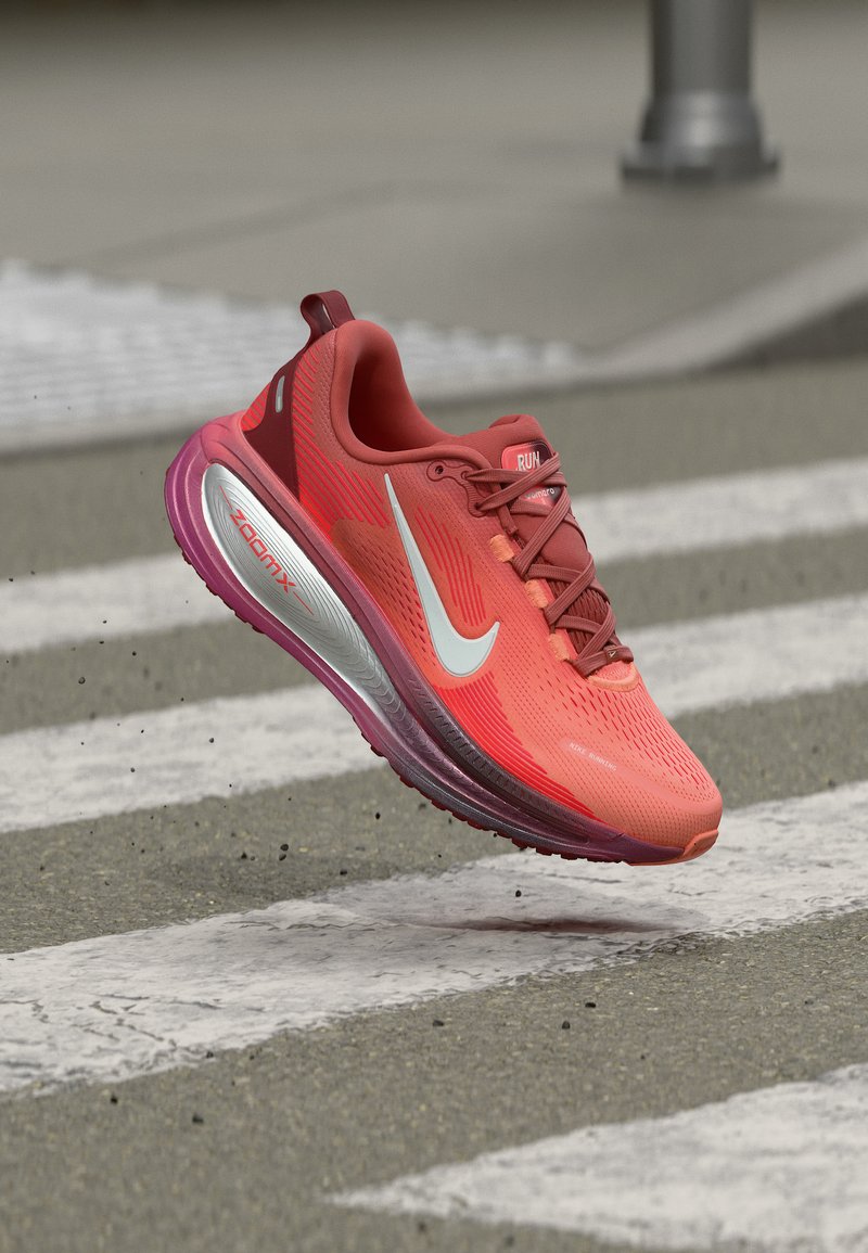 Chaussure de course Nike rouge et violet en plein vol au-dessus d'un passage piéton pavé, mettant en avant la semelle amortissante ZoomX et le logo Swoosh blanc.