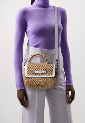 Personne portant un col roulé violet et un pantalon gris clair avec un sac à main en paille tressée doté d'une poignée en bambou et d'une sangle grise.