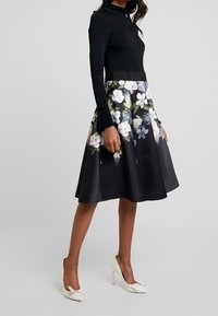 Zwarte lange mouwen top met een gerimpelde halslijn. De rok is knie-lengte, zwart satijn met een bloemenprint in wit, blauw en groen. Witte hoge hakken.