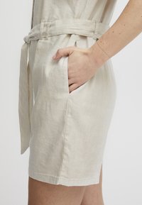 Shorts en lin beige clair avec une taille ceinturée, des poches latérales et une texture lisse, offrant une coupe décontractée au niveau des cuisses.