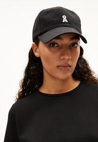 Schwarze Baseballmütze mit gebogenem Schirm, auf der Vorderseite ist ein weißes Logo zu sehen. Tragen von einer Person mit lockigem Haar, die ein schwarzes Oberteil trägt.