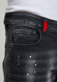 Musta farkut slim fit -mallilla, joissa on punainen etiketti, hienovaraista kulutusta ja valkoisia roiskeita vasemmalla puolella lähellä taskua.