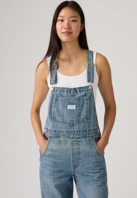 Τζιν φόρμες σε ανοιχτό μπλε χρώμα με ρυθμιζόμενους ιμάντες, μπροστινές τσέπες και μικρό λογότυπο στο στήθος. Το ύφασμα έχει φθαρμένη υφή.