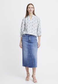 Blouse fleurie blanche avec des manches bouffantes, associée à une jupe crayon en denim bleu taille haute. La tenue est complétée par des sandales blanches à lanières.
