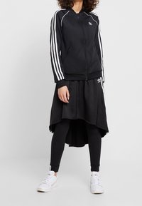 Svart zip-up jacka med vita ränder på ärmarna. Lager av svart kjol över svarta leggings. Vita sneakers med texturerad design.