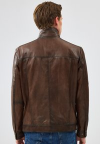Veste en cuir marron avec un col haut, texture lisse et coutures visibles, présentant des poignets à boutons et un design simple et classique.