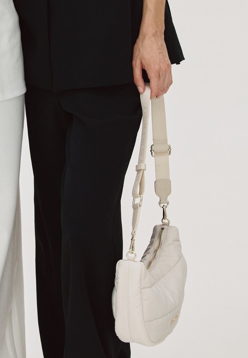 Sac à épaule blanc matelassé avec fermeture éclair, bandoulière réglable et quincaillerie en métal. Le sac est tenu par une personne portant un pantalon noir.
