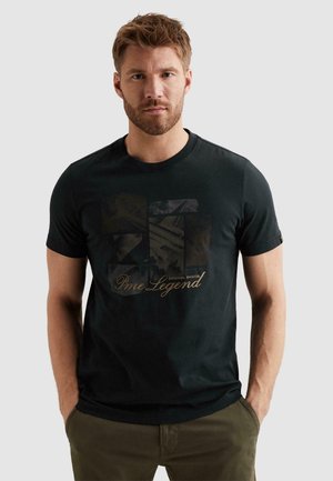Mann mit Bart, der ein schwarzes Grafik-T-Shirt mit dem Schriftzug "Pme Legend" trägt, steht mit den Händen in den Taschen vor einem einfarbigen grauen Hintergrund.