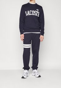 Marinblå sweatshirt med vit "LACOSTE"-logotyp, tillsammans med matchande sweatpants som har vita ränder och logotypdetalj p å höfterna.