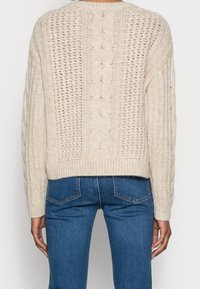 Pull beige clair en maille avec un motif torsadé texturé, dos à tissage ouvert et ourlet côtelé. Associé à un jean en denim bleu.