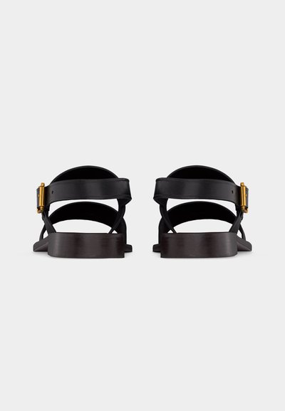Μαύρα δερμάτινα σανδάλια με λουριά στον αστράγαλο και χρυσές αγκράφες, εμφανιζόμενα από πίσω σε απλό ανοιχτό φόντο.
