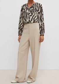 Blouse longue et transparente à manches longues avec un motif abstrait noir et beige, associée à un pantalon large beige. Des chaussures plates beiges complètent le look.