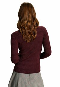 Femme aux longs cheveux bruns ondulés portant un pull texturé bordeaux foncé et une jupe à carreaux grise, vue de dos sur un fond blanc.