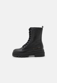 Bota de tobillo de cuero negro con una superficie texturizada, diseño de cordones, suela gruesa y un acento de logo dorado en el costado.