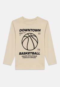 Bēša garām piedurkņu T-kreklā ir melna basketbola grafika, teksts "DOWNTOWN BASKETBALL" un dažādi sporta tematiskie izteicieni.