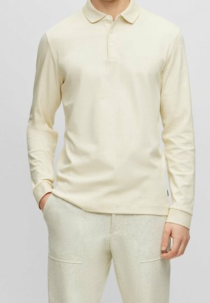 Mężczyzna w kremowej koszulce polo z długim rękawem i jasnobeżowych spodniach, stojący z jedną ręką w kieszeni na jednolitym tle.