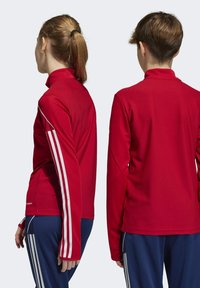 Chemise de sport rouge à manches longues avec un col montant, tissu texturé et accents de rayures blanches sur les manches, associée à un pantalon marine.