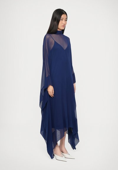 Rochie albastru marin din țesătură transparentă, cu gât înalt, mâneci largi și lungi, tiv asimetric și siluetă drapată. Combinată cu pantofi albi.