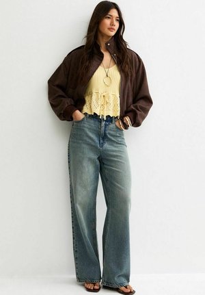 Vrouw draagt een oversized bruine jas, gele gebreide top, wijde broekspijpen jeans, sandalen en armbanden, staat met handen in de zakken tegen een witte muur.