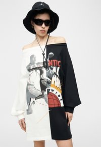 Γυναίκα με υπερμεγέθη ασπρόμαυρ ο φόρεμα έξω από τον ώμο με στάμπα καλαθοσφαιριστή, μαύρο καπέλο τύπου μπαλτά και μαύρα γυαλιά ηλίου.