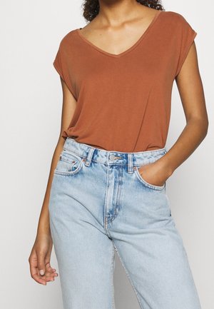 Kobieta ubrana w rdzawy top z krótkim rękawem i dekoltem w serek, włożony do jasnoniebieskich dżinsów z wysokim stanem, z jedną ręką w kieszeni.