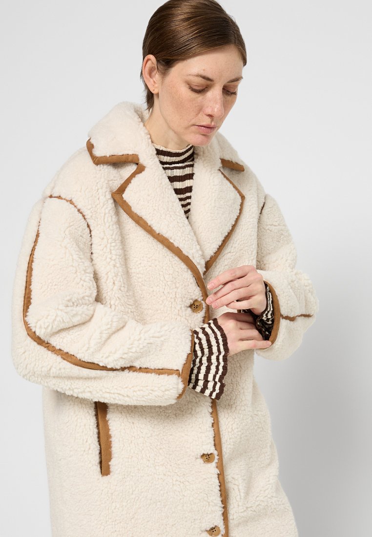 Cremefarbener Fleece-Mantel mit braunen Akzenten, abgestepptem Kragen und Knopfverschluss. Gestreifte Innenmanschetten sichtbar, weiche Textur überall.