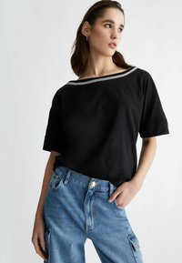 Top nero a maniche corte con ampia scollatura a barca e bordatura argentata, abbinato a jeans cargo in denim blu chiaro.