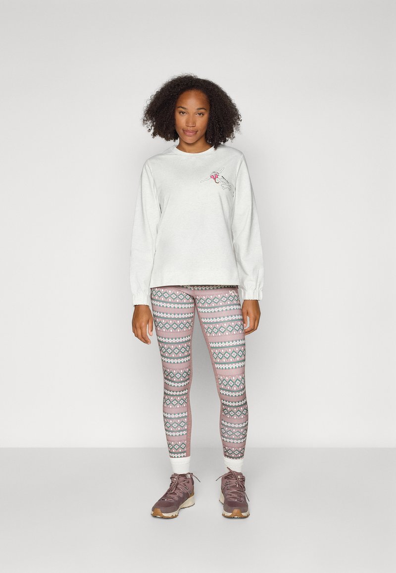 Hellgraues Langarmshirt mit einem kleinen Grafikdesign, kombiniert mit mehrfarbigen gemusterten Leggings, die geometrische Formen zeigen, und Stiefeln.