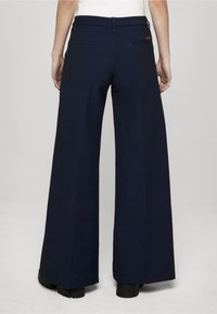 Plaša griezuma tumši zili svārki no teksturēta auduma ar aizmugurējām kabatām un oranžām šuvēm. Jostasvieta ir gluda ar jostas cilpām.