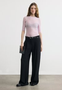 Γυναίκα που φοράει ροζ ανοιχτό ριμπ κοντομάνικο τοπ, φαρδιά μαύρη παντελόνια με ζώνη, κρατώντας μαύρο τσαντάκι σε λευκό φόντο.