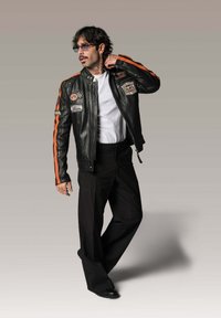Veste de moto en cuir noir avec des rayures orange, des écussons et des fermetures éclair, associée à un pantalon noir et un t-shirt blanc.