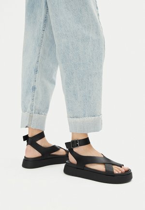 Schwarze Plateau-Sandalen mit breiten, flachen Riemen. Mit einer Schnallenverschluss und einer strukturierten Sohle. Getragen zu hellblauen Jeans mit Umschlag.