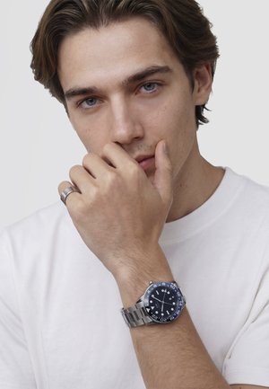 Junger Mann mit blauen Augen, der ein weißes Hemd, einen silbernen Ring und eine Edelstahl-Uhr mit schwarzer und blauer Lünette an seinem linken Handgelenk trägt.