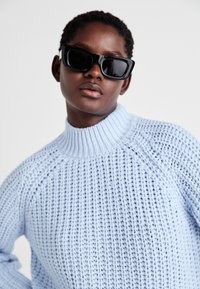 Ανοιχτό μπλε πλεκτό πουλόβερ με ψηλό, ριμπ λαιμό και υφασμάτινη υφή. Το μοντέλο φοράει υπερμεγέθη μαύρα γυαλιά ηλίου.