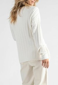 Crème kabelgebreide trui met ribbeldetails, relaxte pasvorm en pofmouwen. Gecombineerd met lichtgekleurde broek. Zachte textuur.
