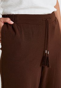 Gros plan sur un pantalon marron texturé avec une taille élastique et un cordon de serrage, comportant des pompons décoratifs aux extrémités.
