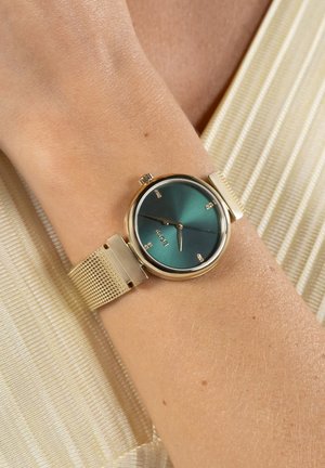 Montre-bracelet à bracelet en maille dorée avec un cadran vert et des marqueurs horaires minimalistes, portée au poignet d'une personne sur un tissu rayé clair.